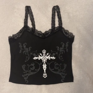 Svart croppad linnetopp med spets och korsmotiv - Säljer en svart croppad linnetopp med spetsdetaljer vid axelband och urringning. Säljer då jag ej använder den längre, minns ej om den är köpt från AliExpress eller SHEIN. Sitter som S/XS