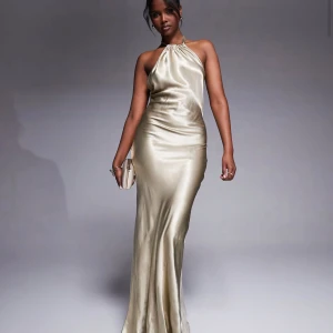 Champagnefärgad långklänning i satin - Säljer en oanvänd elegant champagnefärgad långklänning i satin med halterneck och öppen rygg. Klänningen har knytning i nacken och en figurnära passform. Prislappen sitter kvar 