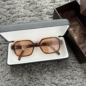 Bruna solglasögon från Corlin Eyewear - Snygga solglasögon från Corlin med bruna, rektangulära bågar och matchande bruna glas. Kommer i originalfodral och ask. 