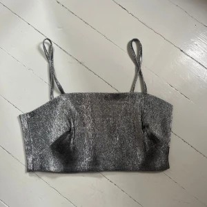 Glittrig silverfärgad croppad linnetopp - Säljer en glittrig silverfärgad croppad linnetopp med smala axelband. Toppen har en stilren och festlig look, perfekt för att sticka ut. Materialet är glansigt och ger en lyxig känsla.