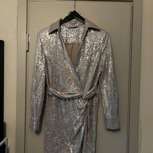 Glittrig silverklänning från Zara - Säljer en glittrig silverfärgad klänning från Zara med omlottmodell och bälte i midjan. Klänningen har lång ärm, krage och är täckt av paljetter som ger en riktigt festlig look. 
