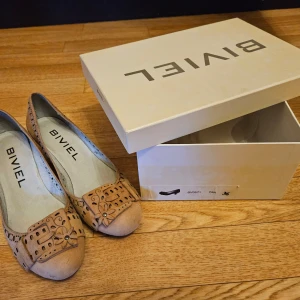 Bruna ballerinaskor från Biviel - Säljer ett par snygga ballerinaskor från Biviel i ljusbrunt skinn med dekorativt mönster och rosett framtill. Skorna har rund tå och fina utskärningar som ger en unik look. Perfekta för dig som vill ha något stilrent men ändå speciellt.