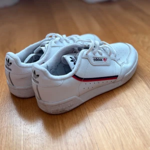 Vita Adidas Continental 80 sneakers - Säljer ett par klassiska vita Adidas Continental 80 sneakers med röd och marinblå rand längs sidan. Skorna har snörning, perforerade detaljer och Adidas-logga på sidan och hälen. Perfekta för en stilren och sportig look.