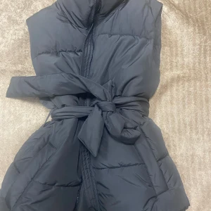 Svart väst med bälte från Lindex - Säljer en svart dunväst från Lindex med hög krage och bälte i midjan. Västen har dragkedja framtill och en stilren. Aldrig använd, utan prislapp 