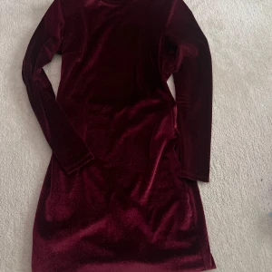 Vinröd sammetsklänning med öppen rygg - Säljer en vinröd sammetsklänning med lång ärm och glittrig finish. Klänningen har en snygg öppen rygg och är figurnära i modellen. Perfekt för dig som vill sticka ut lite extra!