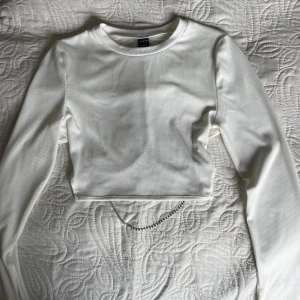 Vit croppad långärmad topp med öppen rygg - Säljer en vit croppad långärmad topp i ribbat material. Toppen har en tight passform och är dekorerad med silvriga kedjedetaljer på baksidan som ger en unik look. Perfekt för dig som gillar trendiga plagg! Storlek XS