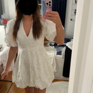 Vit klänning - Superfin vit kortklänning med puffiga kortärmar och v-ringning. Klänningen har ett subtilt blommigt mönster i tyget och en lätt volang i nederkanten. Perfekt för sommarens alla tillfällen!