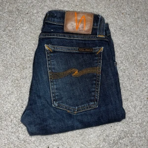 Mörkblå jeans från Nudie Jeans - Säljer ett par mörkblå jeans från Nudie Jeans med klassisk femficksdesign och orangea sömmar. Jeansen har en slim fit och är tillverkade i denim med snygga detaljer på bakfickan och läderpatch i midjan.