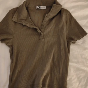 Grön ribbad pikétröja från FB Sister - Säljer en oliv grönribbad pikétröja från FB Sister i storlek XS. Tröjan har korta ärmar, klassisk krage och knappar framtill. Använd fåtal gånger. Den är croppad