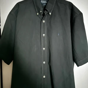Polo Ralph Lauren - Klassisk svart skjorta från Polo Ralph Lauren med korta ärmar och knappar framtill. Skjortan har button-down krage och diskret broderad logga på bröstet. Tillverkad i 100% bomull för en skön känsla. M storlek passar även L bra