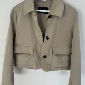 Beige croppad jacka från Massimo dutti  - Snygg beige croppad jacka med stora fickor framtill och tre bruna knappar. Jackan har klassisk krage och lång ärm, perfekt för vår och höst. Enkel och stilren design som passar till mycket.