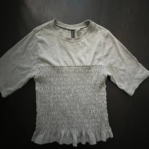 Grå topp med smock och volang från H&M Divided - Säljer en ljusgrå topp från H&M Divided med smockad midja och volangkant nedtill. Toppen har korta ärmar och rund halsringning. Perfekt för en avslappnad och trendig look.