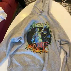 Grå Metallica hoodie med tryck - Säljer en grå hoodie från Metallica med stort färgglatt tryck av Lady Justice och texten 'And Justice For All' på framsidan. Baksidan har låtlista i blått och gult. Perfekt för dig som gillar bandmerch och vill sticka ut.