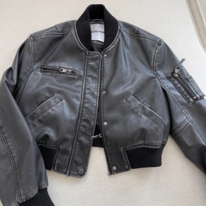 Svart croppad skinnjacka från Bershka - Säljer en svart croppad skinnjacka från Bershka med ribbade muddar och dragkedjedetaljer. Jackan har fickor med lock och dragkedja samt en cool ficka på ärmen. Perfekt för dig som vill ha en edgy stil.