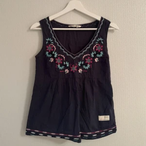 Mörkblå broderad linnetopp från Odd Molly - Supersöt mörkblå linnetopp från Odd Molly med v-ringning och broderade blommor i rosa, turkos och vitt framtill. Toppen är ärmlös och har dekorativa detaljer längs nederkanten. Perfekt för varma dagar!