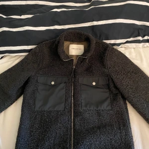 These Glory Days jacka - Säljer en svart teddyjacka från These Glory Days i storlek small. Jackan har två stora fickor framtill med lock och knappar samt dragkedja. Mjuk och mysig med en modern look, perfekt för kyliga dagar.