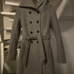 Grå kappa med huva och bälte - Säljer en stilren grå kappa med dubbelknäppning, huva och knytskärp i midjan. Jackan har långa ärmar och en klassisk, figurnära passform. Perfekt för kyliga dagar när du vill vara både varm och snygg. Innehåller bland annat  ull.  Storlek S  Nypris var  1400kr