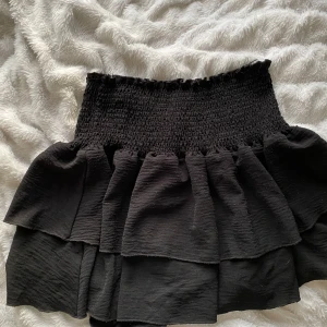 Svart volangkjol från Skue - Supersöt svart kjol från Skue med bred smock i midjan och dubbla volanger. Kjolen är kort och har en luftig känsla, perfekt att styla med en enkel topp. Storlek S (EU 36).