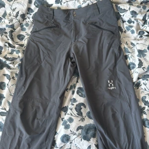 Grå friluftsbyxor från Haglöfs - Säljer ett par grå friluftsbyxor från Haglöfs med dragkedjor vid fickorna och logga på ena benet. Byxorna har normal passform och är perfekta för utomhusaktiviteter. Snygg och funktionell design med praktiska detaljer.