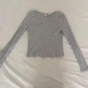 Grå ribbad långärmad topp  - Säljer en ljusgrå ribbad långärmad topp från H&M. Toppen har spetsdetaljer och en liten rosett vid halsringningen. Perfekt för lager-på-lager och enkel att matcha med olika outfits.