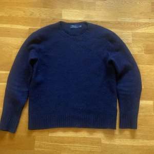 Kashmirtröja från Polo Ralph Lauren - Säljer en klassisk mörkblå tröja från Polo Ralph Lauren i ull och kashmir blandning. Tröjan har rund halsringning och är perfekt för kyligare dagar men även fina vårdagar samt finare tillställningar. Enkel och stilren design som passar till det mesta. Priset är inte hugget i sten👍 hör av dig vid frågor