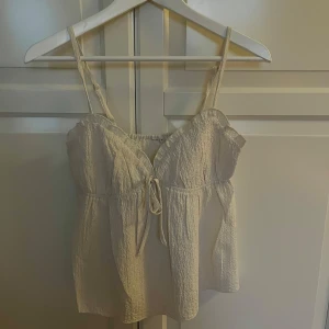 Beige linnetopp med knytning - Säljer en ljusbeige linnetopp med smala axelband och knytning framtill. Toppen har en lätt rynkad struktur och är tillverkad i ett tunt, luftigt material. Perfekt för varma dagar och enkel att matcha med olika outfits.