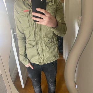Olivgrön fieldjacka med fickor - Säljer en olivgrön fieldjacka från Superdry med flera praktiska fickor och dragkedja framtill. Jackan har en klassisk militärinspirerad look och är perfekt för våren/hösten. Jackan har en liten defekt på högerfickan som inte alls syns vid användning! Pris är ej hugget i sten!!!🪨