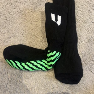 Svarta sportstrumpor med grönt greppmönster - Säljer ett par svarta sportstrumpor från Unisport med vitt logomärke och grönt greppmönster undertill. Perfekta för dig som vill ha extra grepp i skorna under träning eller match. Helt oanvända. 