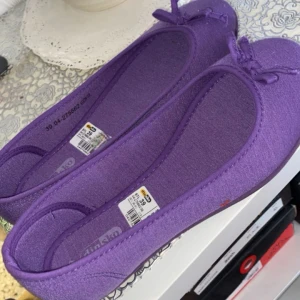 Lila ballerinaskor med rosett från Din Sko - Säljer ett par lila ballerinaskor från Din Sko. Skorna har en söt liten rosett framtill och är tillverkade i ett mjukt syntetmaterial. Perfekta för dig som vill ha ett par lätta och bekväma skor i en härlig färg.