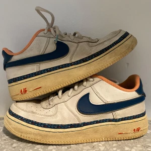 Nike Air Force 1 med blå och orange detaljer - Nike Air Force 1 sneakers i vitt med blå swoosh och orange insida. Skorna har en beige sula med färgade detaljer och klassisk snörning. 