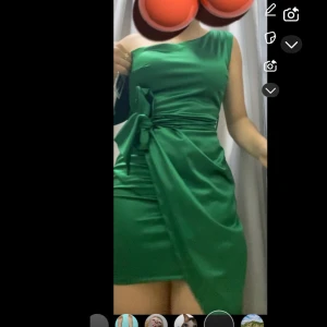 Grön oneshoulder kort klänning med knytband - Säljer en smaragdgrön kortklänning i satin med oneshoulder-design och brett knytband i midjan. Klänningen har en omlottkänsla och elegant drapering framtill. Perfekt för dig som vill sticka ut med en trendig och stilren look.