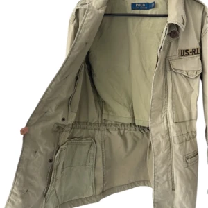 Army jacka från Polo Ralph Lauren - Säljer en army jacka från Polo Ralph Lauren med flera fickor framtill, axelklaffar och broderad detalj 'US-RL' på bröstet. Jackan har knäppning med knappar och klassisk krage. Perfekt för dig som gillar en stilren och funktionell look. Storlek S. Borde passa om man har M i vissa fall.