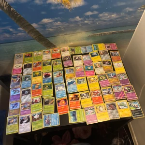 Samling Pokémonkort - Stor samling Pokémonkort med blandade motiv och färger. Kortleken innehåller flera olika Pokémon, inklusive Rillaboom som syns överst i högen. Perfekt för samlare eller för dig som vill utöka din kortlek. Kort i standardstorlek, hålls ihop med ett blått gummiband.