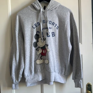 Grå hoodie med Mickey Mouse från Divided x Disney - Grå hoodie med tryck av Mickey Mouse och texten 'Les Sports Club' framtill. Tröjan har huva med dragsko och ribbade muddar vid ärmslut och nederkant. Perfekt för en avslappnad stil.
