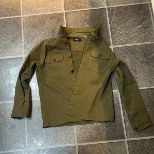 Olivgrön skjorta från Gabba - Passar M!!! Snygg olivgrön skjorta från Gabba med två bröstfickor och knappar framtill. Jag säljer då jag inte använder den längre! Helt ny skick och knappt använd!!