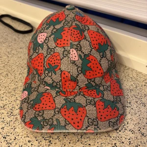 Gucci keps med jordgubbsmönster - Cool keps från Gucci med färgglatt jordgubbsmönster på grå botten och klassiska GG-loggor. Justerbar rem baktill i rosa läder. Perfekt statement-piece för dig som gillar unika accessoarer.