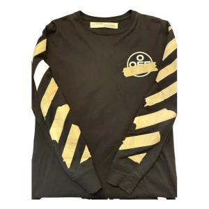 Svart långärmad tröja från Off-White - Svart långärmad tröja från Off-White med ikoniska beigea tejp-liknande ränder på ärmarna och stort korsmönster på ryggen. Rund halsringning och tryckt Off-White-logga framtill. Perfekt för dig som gillar streetwear och vill sticka ut.