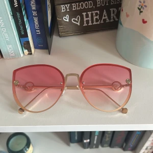 Fendi Rosa solglasögon med guldiga bågar - Trendiga solglasögon med stora, runda linser i rosa ton och tunna guldiga metallbågar. Snygg detalj med cirklar på sidorna och en modern, oversized look. Perfekta för att sticka ut och ge en färgklick till din outfit.