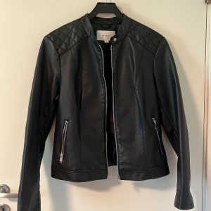 Svart skinnjacka från VILA - Snygg svart skinnjacka från VILA med quiltade detaljer på axlar och nedtill. Jackan har dragkedjor framtill och vid fickorna samt en stilren, kraglös design. Lite tecken på användning längst ut på ärmarna, annars ser den som ny ut!