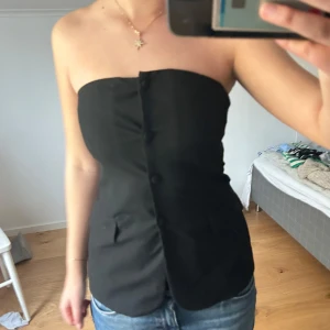 Svart tubtopp med knappar - Snygg svart tubtopp med knappar framtill och fickdetaljer. Stilren och enkel design som passar till mycket. Perfekt att styla med jeans eller kjol.