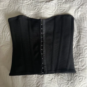 Svart korsett topp - Säljer en svart korsett med hyskknäppning framtill. Modellen är enkel och stilren med markerade sömmar och förstärkningar för extra stöd. Perfekt att styla till olika outfits. Den är storlek 34 och lite liten i storleken men har lite stretch. Aldrig använd.