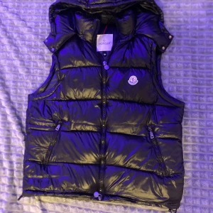  Moncler väst - Säljer en svart glansig dunväst från Moncler med huva och dragkedja framtill. Västen har två snedställda fickor med dragkedja och Moncler-logga på bröstet. Perfekt för lager-på-lager och snygg streetstil. Storlek är XL men passar till L också 