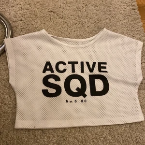 Vit croppad mesh-topp med tryck - Säljer en vit croppad topp/magtröja i meshmaterial med svart texttryck 'ACTIVE SQD' framtill. Perfekt för träning eller sportiga outfits. Tunt och luftigt tyg som andas.