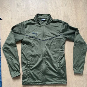 Grön träningströja från Puma - Snygg grön vindjacka från Puma med dragkedja framtill och diskret logga på bröstet. Jackan har hög krage, långa ärmar och två fickor på sidorna. Perfekt för dig som gillar sportig stil och vill ha något lätt att slänga på sig.