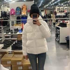 Vit pufferjacka med huva - Säljer en riktigt snygg vit pufferjacka med hög krage och huva. Jackan har en clean look med dragkedja framtill. Liten sminkfläck vid kragen 