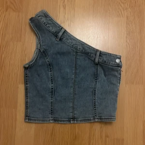 Jeans one shoulder topp från H&M - Cool jeansfärgad topp från H&M i one shoulder-modell med bältdetaljer och dragkedja i sidan. Gjord i denim med synliga sömmar och knappdetalj upptill. Perfekt för dig som vill sticka ut med en edgy look.LITEN I STORLEK!
