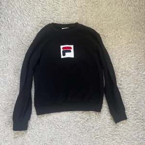Svart sweatshirt från FILA med logga - Svart sweatshirt från FILA i bomull med klassisk rund hals och stor broderad logga framtill i vitt, rött och blått. Tröjan har långa ärmar och ribbade muddar vid ärmslut och nederkant. Perfekt för en sportig och avslappnad stil.