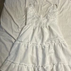 Vit spetsklänning från Designbyamor XS - Supersöt vit kortklänning från Designbyamor i storlek XS. Klänningen har spets upptill, v-ringning, volanger och en stor rosett i ryggen. Perfekt för sommarens alla tillfällen och ger en romantisk vibe med sina fina detaljer.