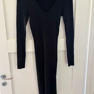 Svart ribbad långklänning - Svart långklänning med ribbad struktur och v-ringning. Klänningen har lång ärm och är figurnära i modellen, perfekt för dig som gillar en stilren och enkel look. Materialet är stickat och stretchigt för en bekväm passform.