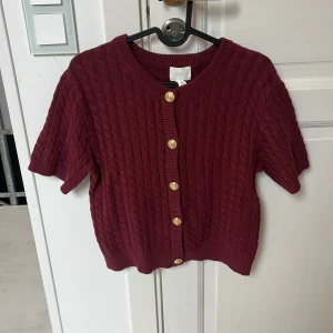 Vinröd kofta med guldknappar H&M - Kortärmad vinröd kofta från H&M med snygg kabelstickad struktur och sex guldiga knappar framtill. Tröjan har rund halsringning och ribbade kanter, perfekt för lager-på-lager och en trendig look.